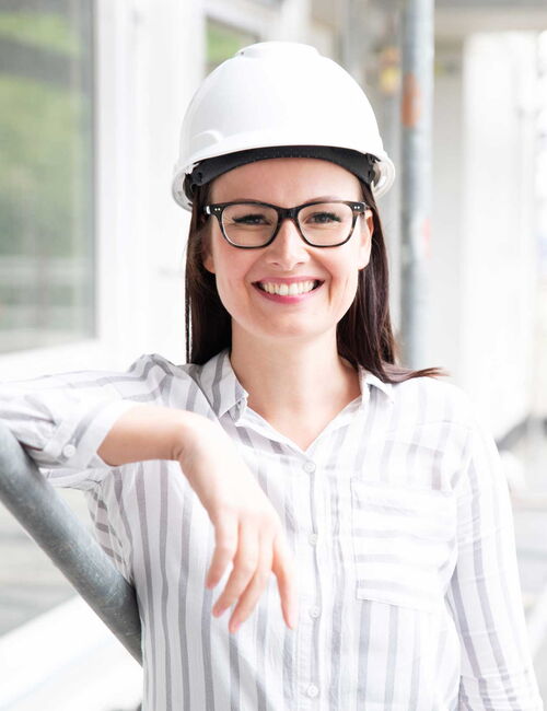Eine Frau lehnt an einer Wand, sie trägt einen weißen Schutzhelm und lächelt.