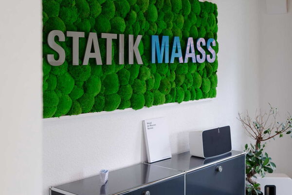 Moosbild mit dem Statik Maass Schriftzug hängt an einer Wand mit moderner Büroeinrichtung.