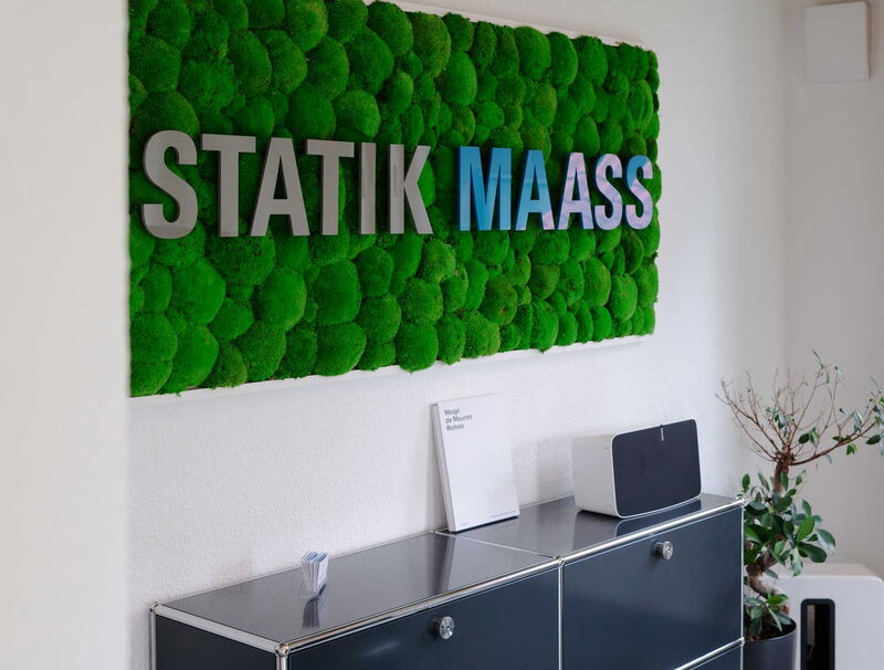 Moosbild mit dem Statik Maass Schriftzug hängt an einer Wand mit moderner Büroeinrichtung.