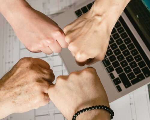 Vier Fäuste stoßen in der Mitte zusammen über einem Laptop und technischen Zeichnungen.