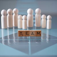Holzbausteine bilden das Wort Team, im Hintergrund stehen weiße Spielpüppchen auf einem Glastisch.