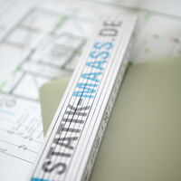 Ein Zollstock mit dem Logoaufdruck von Statik Maaß liegt auf Bauplänen.