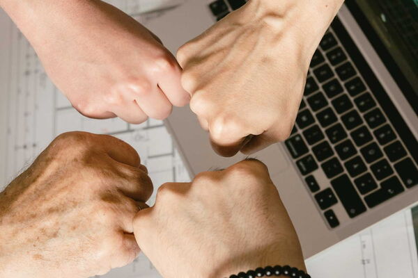 Vier Fäuste stoßen in der Mitte zusammen über einem Laptop und technischen Zeichnungen.