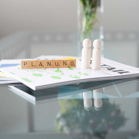 Aus Holzbausteinen wurde das Wort Planung gelegt, im Hintergrund links ist ein Blumenstrauss zu sehen.