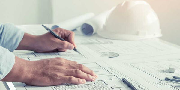 Zwei Hände von links notieren etwas auf einem Bauplan. Im Hintergrund liegt ein Helm.