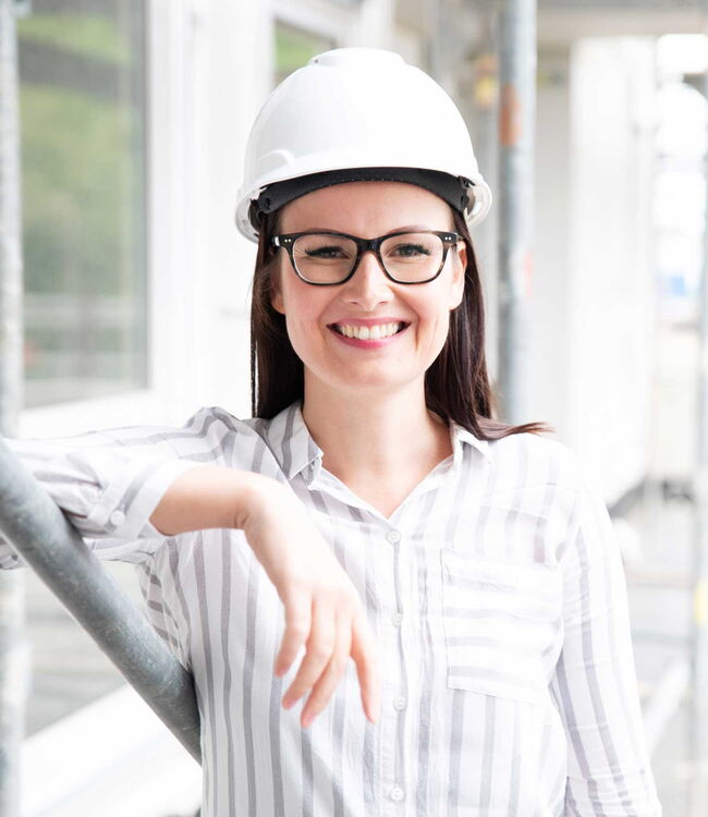 Eine Frau lehnt an einer Wand, sie trägt einen weißen Schutzhelm und lächelt.