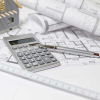 Oberfläche eines Tisches auf welchem Baupläne, ein Taschenrechner und ein Stift liegen, sowie ein kleines Modelhaus steht.